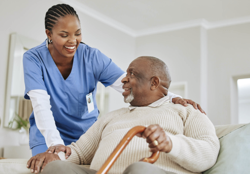 A Black caregiver in blue scrubs smiling with an older man during an in-home care visit.